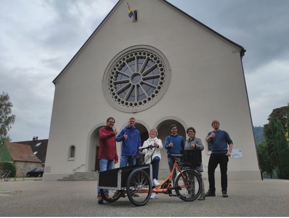 Sechs Menschen, die den Daumen hoch zeigen, vor ihnen ein Lastenrad und hinter ihnen die Dreifaltigkeitskirche.