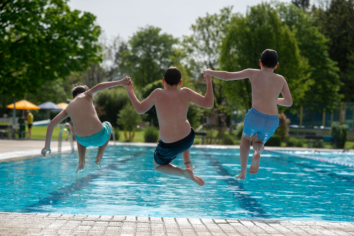 Drei Jungs halten sich an den Händen und springen ins Becken eines Freibads