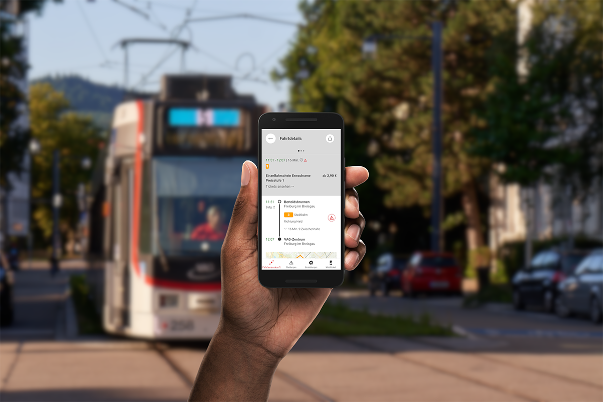 Eine Hand hält ein Handy in die Luft. Darauf zu sehen ist die VAG App. Im Hintergrund fährt eine Straßenbahn der Linie 5. 