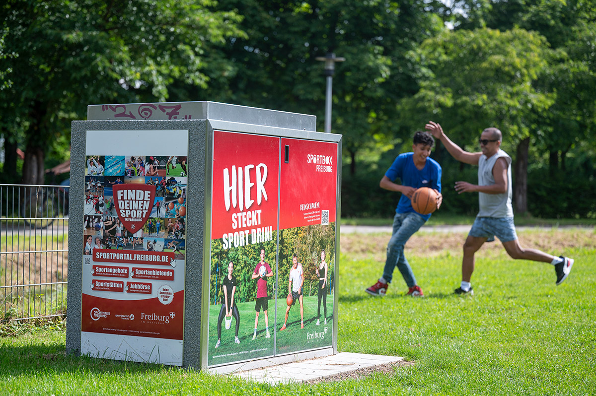 Sportbox, im Hintergrund zwei Basketball spielende Menschen