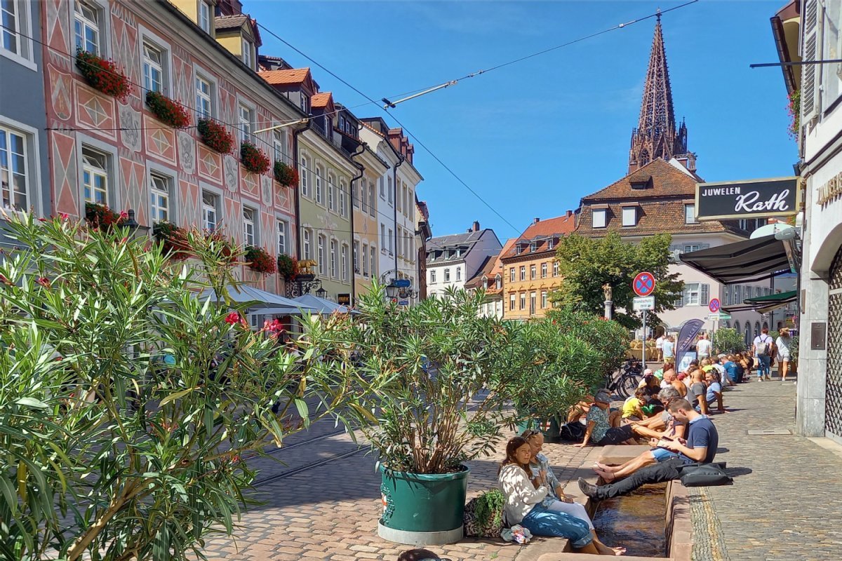 Aufenthaltsqualität in der Freiburger Innenstadt, Salzstraße, Foto: Sibylle Spillmann Historische Häuser mit Münster und gepflasterte Straße und Pflanzkübeln, Menschen sitzen mit den Füßen im